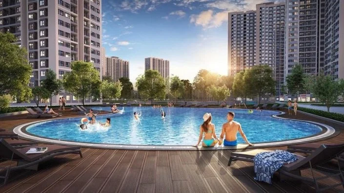 Bể bơi ngoài trời rộng lớn với nhiều người đang bơi lội và thư giãn, xung quanh là các tòa nhà căn hộ