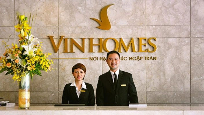 Hai nhân viên lễ tân của Vinhomes, một nam và một nữ, đang đứng sau quầy tiếp tân với logo Vinhomes trên tường.